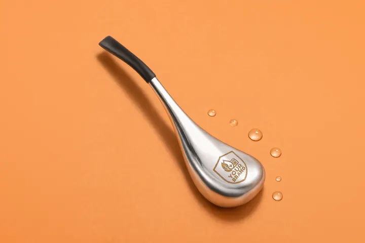 Brushed steel cryo comfort tool with a black grip and a gold YOUR BRAND shield on its head, cold beads of water beside it on peach